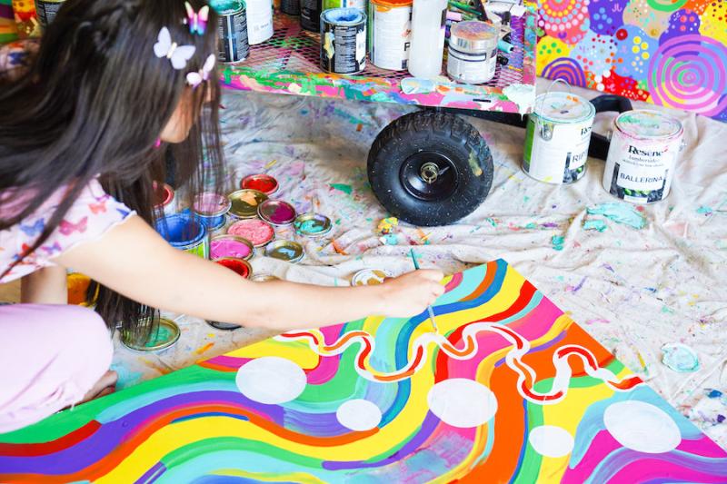 Image is taken over the shoulder of artist Mali Isabel as she sits on the ground looking over one of her colourful artworks, surrounded by colourful paints and brushes