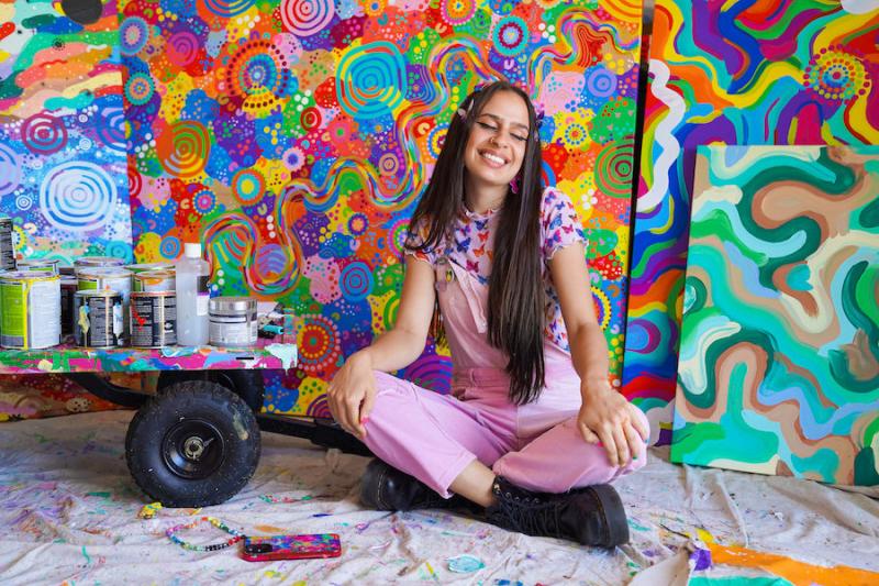 Artist Mali Isabel sits among her colourful artwork with a smile and eyes closed