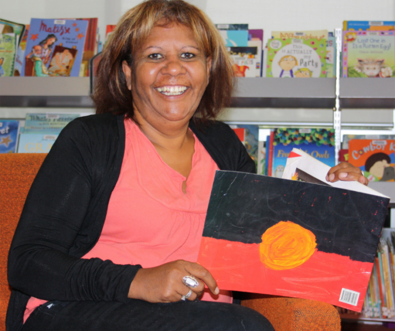 Picture of an indigenous woman wearing a pink top and black cardigan sitting in a library holding a book with the Aboriginal flag on the back